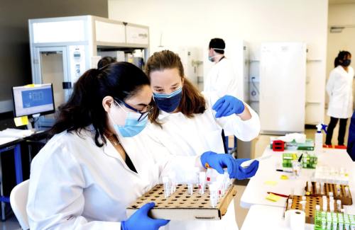 Two scientists wearing gloves and masks place small vials containing COVID-19 test samples into rows.