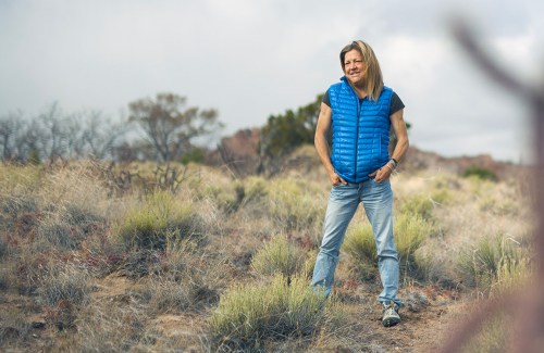 A woman wearing blue jeans and a puffy vest smiles and stands in a field with her hands in her pockets.