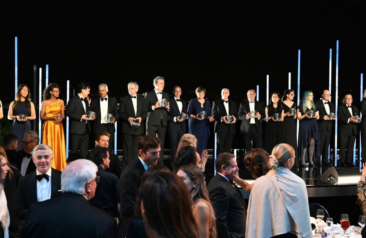 A large group of people stand onstage holding a spherical award in front of a crowd at the 9th annual Breakthrough Prize ceremony