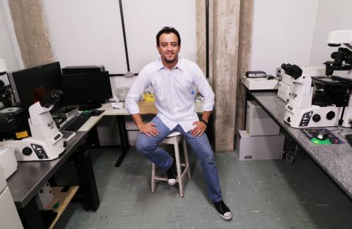 Biologist Gustavo B. Menezes of Brazil sits on a stool in a lab, with microscopes and computer screens.