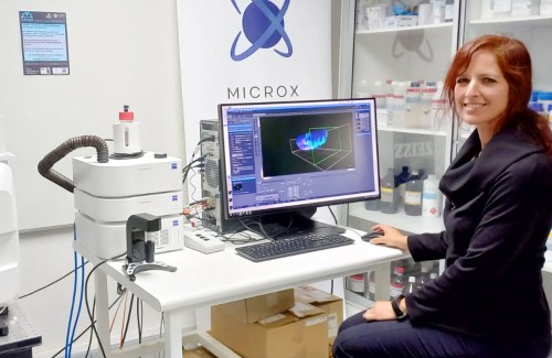 Alenka Lovy smiling in a lab, posing in front of a computer with imaging software open next to a light-sheet microscope.