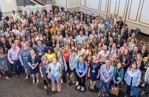 A large, diverse group of people, all wearing conference badges, are gathered together in a ballroom, smiling and looking up at the camera for a group photo.