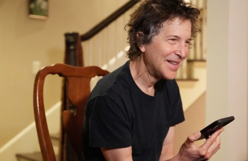 David Baker in a black t-shirt, sitting in a dining chair, smiling with his cell phone in his hand.