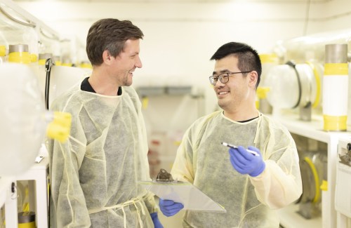 Scientist Noah Palm stands next to a lab member dressed in yellow PPE gowns and blue gloves. They smile at each other. A lab is seen in the background.