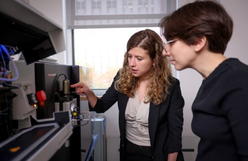 Two women stand side-by-side and look at a piece of lab equipment. One of the women points something out to the other.