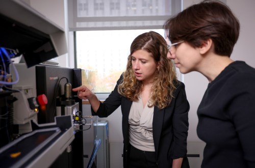 Two women stand side-by-side and look at a piece of lab equipment. One of the women points something out to the other.