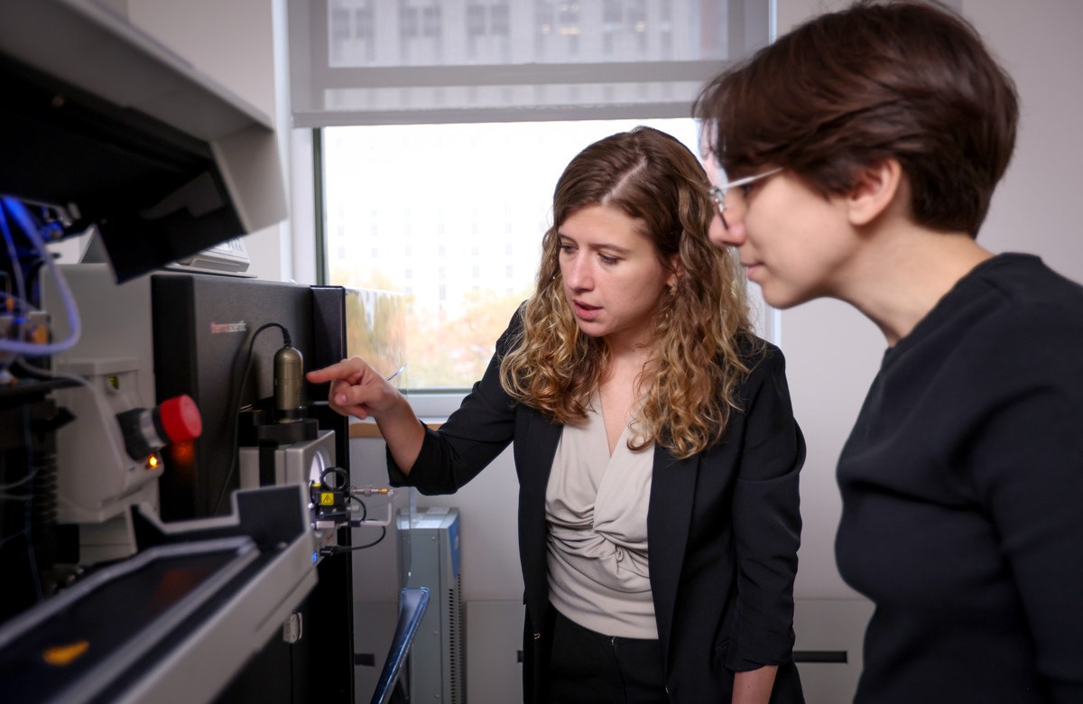 Two women stand side-by-side and look at a piece of lab equipment. One of the women points something out to the other.