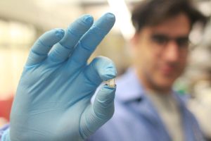 Gloved hand holding a small medical sensor in a lab, researcher blurred in the background