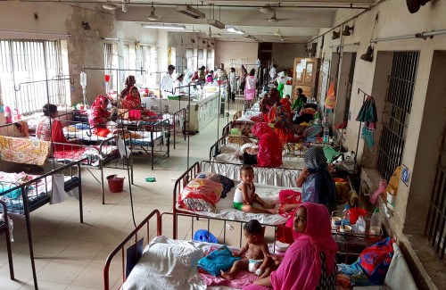 A crowded hospital ward with women and children sharing beds and receiving care. Nurses and doctors are visible in the background tending to patients.