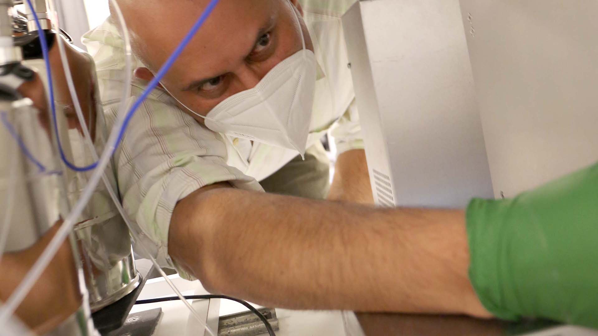 A man wearing a mask works to fix a machine in a lab. A blue wire hangs in front of his face.